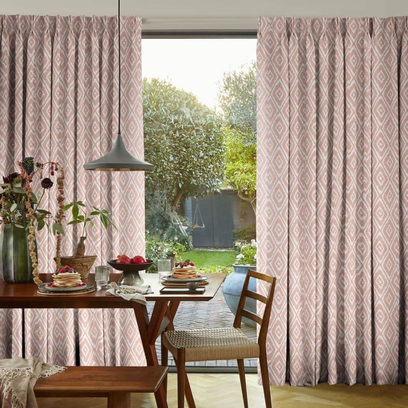 Pleated pink diamond-pattern curtains, drawn apart over a sliding glass door; filtering soft daylight into a dining room with wooden table, pendant lamp and a sunlit garden view.