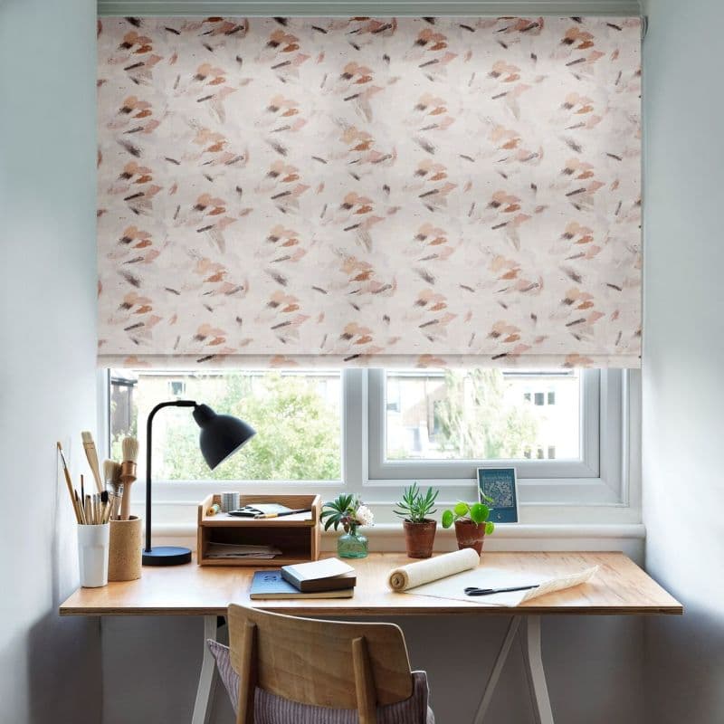 A patterned roller blind (soft pink‑beige brushstrokes) is lowered over the window, diffusing daylight; below, a wooden desk holds a lamp, art brushes, notebooks and potted plants, with trees outside.