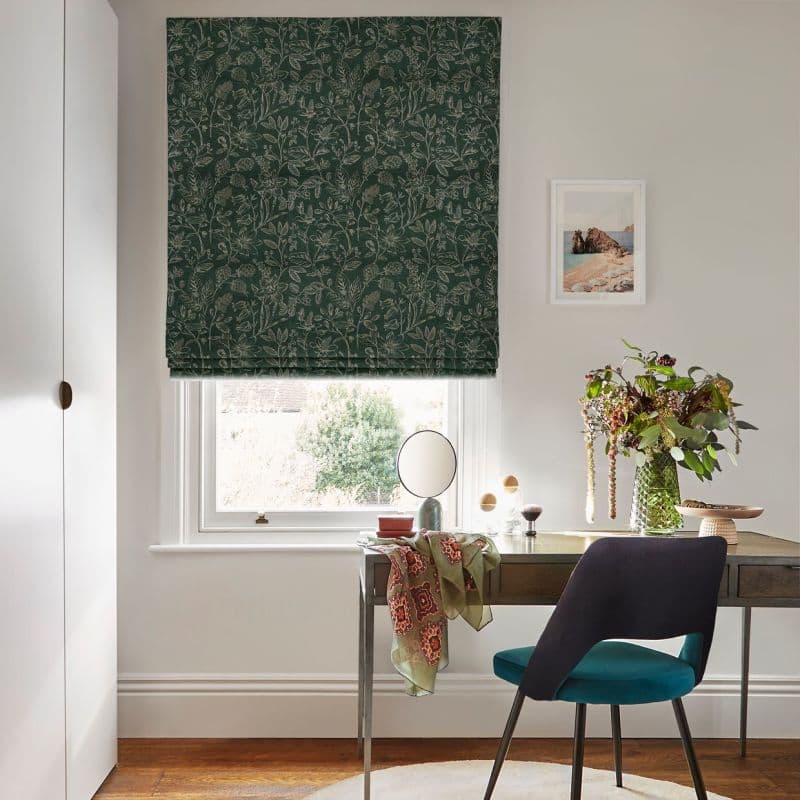 Dark green floral Roman blind, lowered over the window, patterned with pale botanical motifs; filtering daylight into a bright home office with desk, chair, vase of flowers, and framed seaside print.