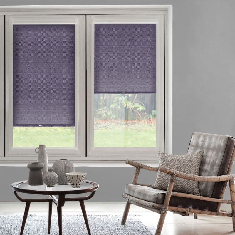 Purple honeycomb cellular blinds on two adjacent window panes—left fully lowered, right halfway raised—filter daylight and obscure a backyard view in a softly lit gray living room with rattan armchair.