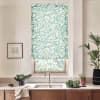 Teal leaf-patterned roller blind covers a kitchen window above a sink; stainless faucet centered on a wood countertop with bowls of greens, potted plants, dish towel, and framed art.