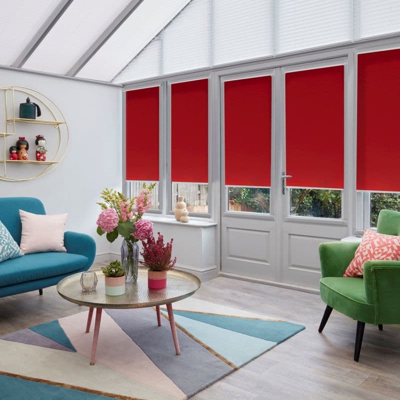 Red roller blinds covering four glazed panels and a door, lowered to mid-height; blocking light while leaving narrow lower panes visible — bright conservatory with pitched glass roof, colorful seating.