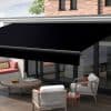 Black retractable cassette awning, fully extended and angled, shading a patio; covers glass sliding doors above wood decking, with outdoor seating, low table, and daytime soft natural light.