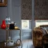 Textured dark grey Roman blinds, lowered over two tall windows with red trim, partially blocking light; in a living room with grey paneled walls, patterned sofa, wooden floor, garden visible below.