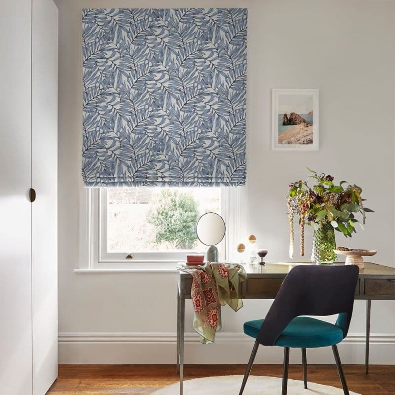 Blue-and-white leafy Roman blind lowered over a window, gently filtering daylight; a desk beneath holds a mirror, vase of flowers and accessories beside a teal chair in a bright home office.