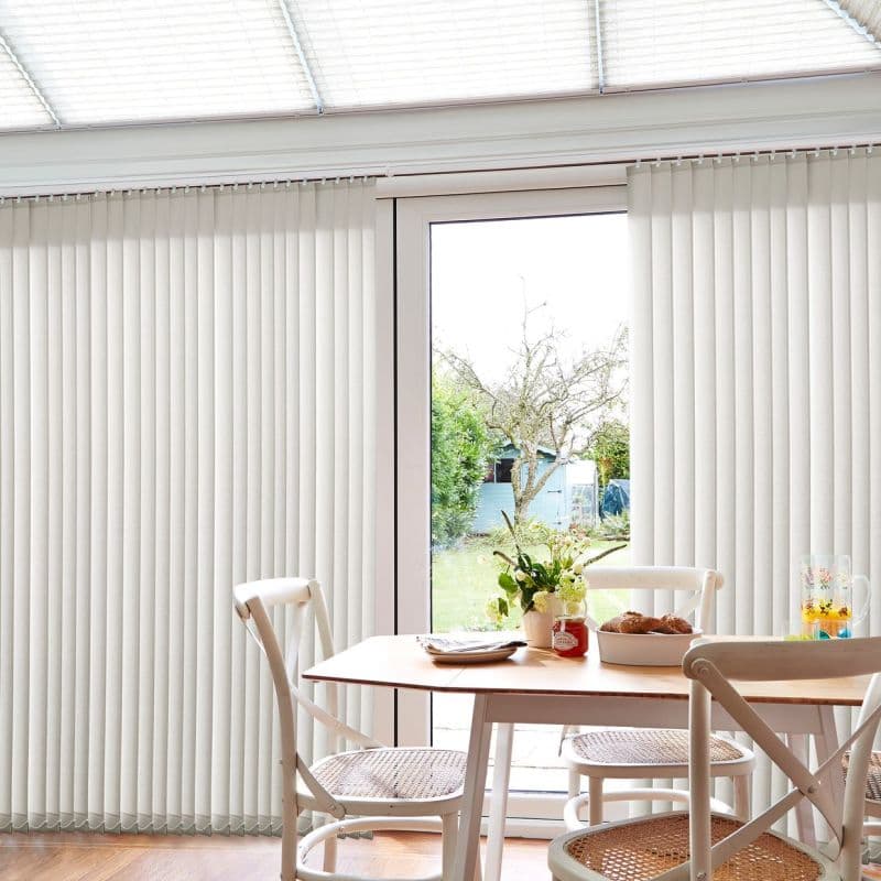 Fabric vertical blinds, floor-to-ceiling and mostly closed with a central gap, filtering bright daylight; dining table and chairs in a sunlit conservatory opening onto a grassy backyard.