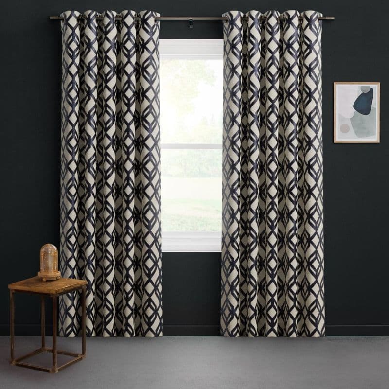 Grommet-top floor-length curtains with navy lattice pattern on cream, hung on a metal rod and parted to reveal a sunlit window, framing a bright green yard against dark-painted walls.