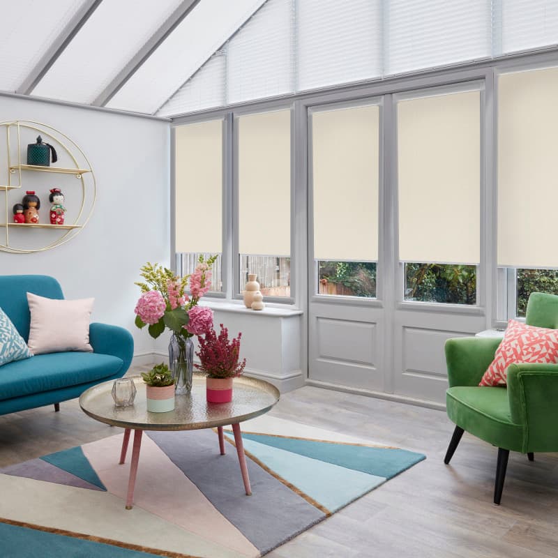 Beige fabric roller blinds lowered two-thirds over tall glass doors and windows, with white pleated blinds in the pitched roof, filtering soft daylight into a colorful conservatory-style living room.