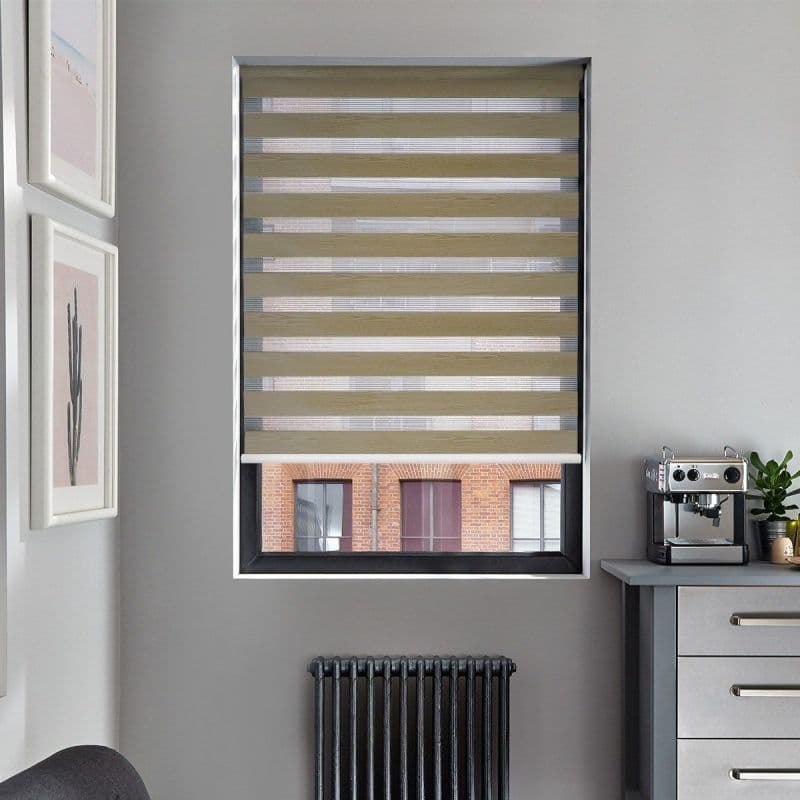 Zebra roller blind, beige alternating opaque-and-sheer horizontal bands, lowered across a recessed window; filtering light, partially revealing a brick building, in a gray kitchen corner with radiator and espresso machine.
