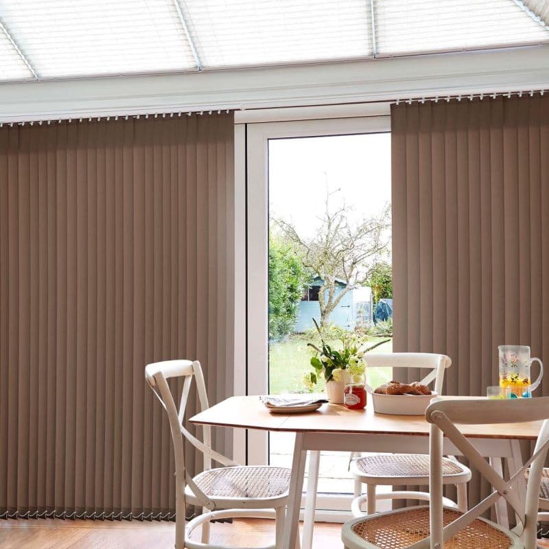 Beige vertical slatted blinds, hung floor-to-ceiling over a patio door, are parted centrally, filtering daylight into a dining nook with a wood table, white chairs, vase and backyard view.