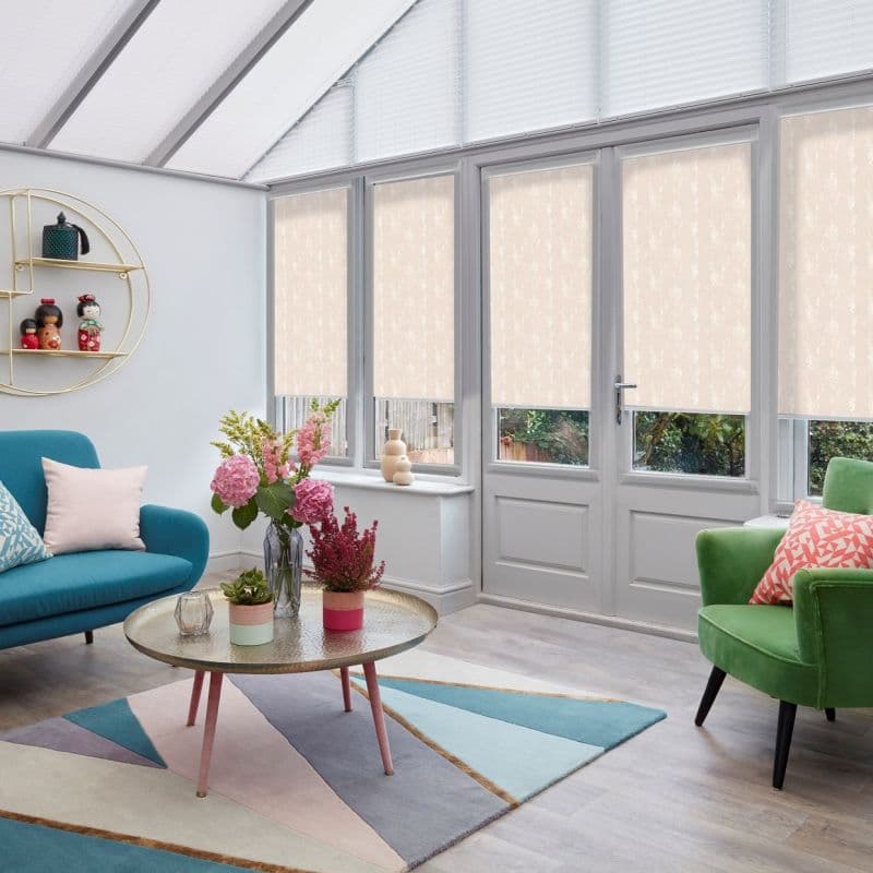 Beige patterned roller blinds lowered over tall glass doors and windows, filtering soft daylight; conservatory-style living room with glass roof, teal sofa, green armchair, round coffee table and floral arrangements.