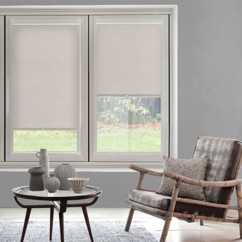 Beige fabric roller blinds, inside-mounted across three windows and partially lowered (center blind slightly raised), filtering daylight; in grey-walled living room with rattan armchair, round coffee table and lawn view.