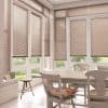 Beige horizontal blinds covering tall French doors and windows, lowered with slats mostly closed and softly filtering daylight; sunlit dining corner with wooden table, chairs, floral cushions and garden view.