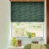 Green patterned Roman blind lowered in soft folds, partly blocking and filtering daylight, in a bedroom window over a sill with glass vases, a potted plant, small fan and green bed.