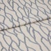 Beige fabric with repeating blue rope-like chain pattern, folded and layered across itself; close-up on a flat surface with soft, even lighting that emphasizes the woven texture.
