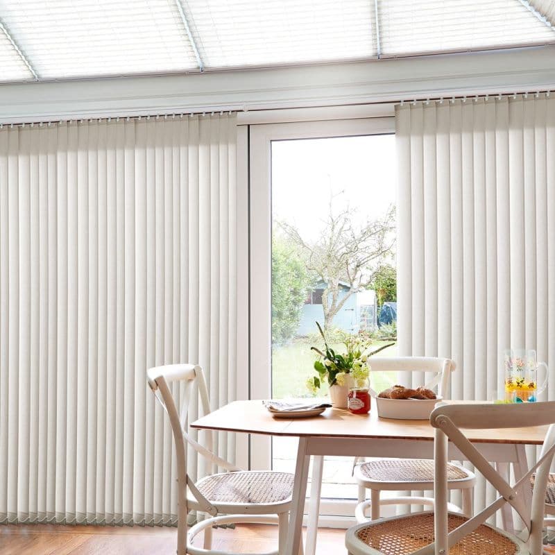 Beige vertical blinds covering a sliding glass door, partly drawn to reveal a central opening; filtering soft daylight into a small dining area with wooden table, chairs, and garden view.
