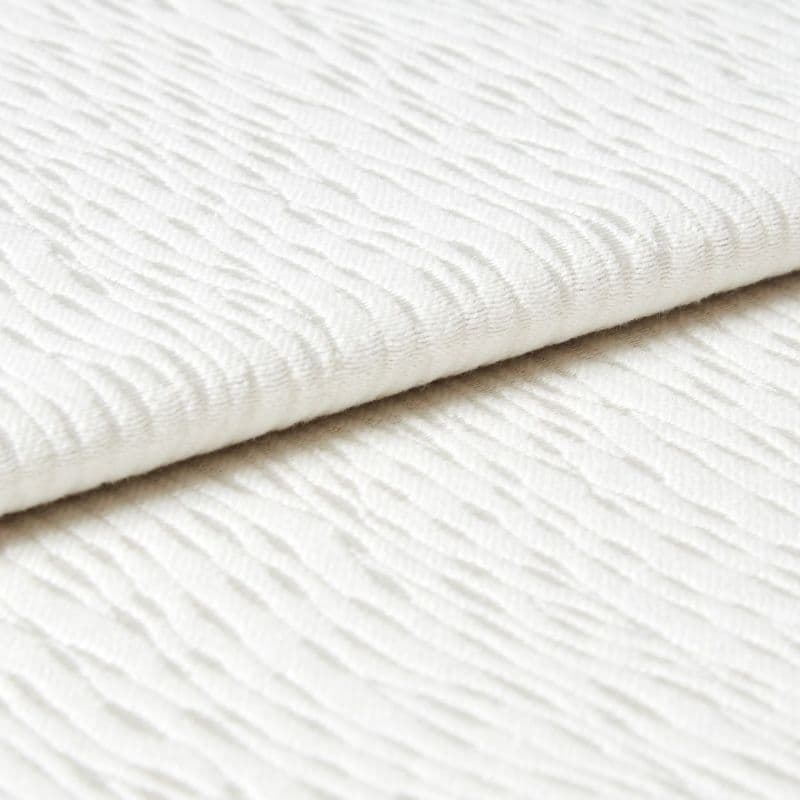 White wavy-ribbed fabric folded with an overlapping edge; fabric lies flat and layered. Close-up view under soft, diffuse light on a neutral surface, suggesting bedding or upholstery.