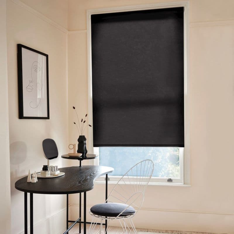 Black roller blind, lowered over a tall window, smooth blackout fabric with a faint horizontal crease; blocking most light. Context: minimalist pale-walled workspace with dark curved desk, wire chair, framed line-art.