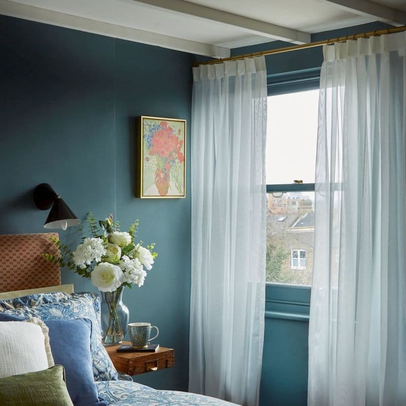 Sheer white curtains on a brass rod, drawn to frame a window and filtering daylight; bedroom with teal walls, patterned bedding, nightstand holding a vase of white flowers and a rooftop view.