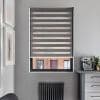 Gray zebra roller blind, lowered over window with alternating opaque and sheer bands, filtering daylight; framed artwork left, black radiator below, countertop with espresso machine to right in gray room.