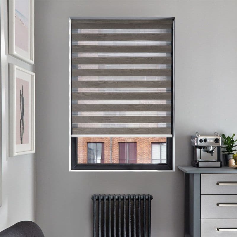 Gray zebra roller blind, lowered over window with alternating opaque and sheer bands, filtering daylight; framed artwork left, black radiator below, countertop with espresso machine to right in gray room.