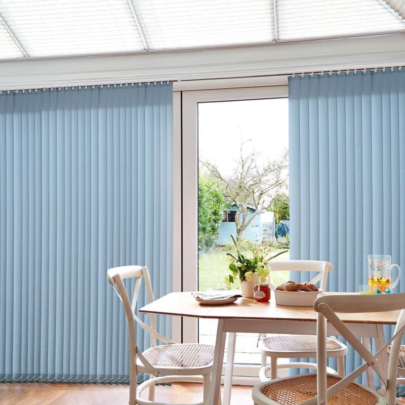 Light-blue vertical blinds cover a sliding glass door, panels mostly closed with a central opening; they partly block light, revealing a sunlit garden and a small dining table with chairs.