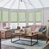Pale green pleated cellular blinds, lowered over tall bay windows, filtering soft daylight; conservatory living room with beige sofa draped by a chunky pink throw, wooden coffee table and rug.