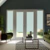 White double glass doors with lowered blinds, framed by sidelights, stand closed, admitting filtered light into a living room with green armchairs, glass coffee table and zigzag rug; garden beyond.
