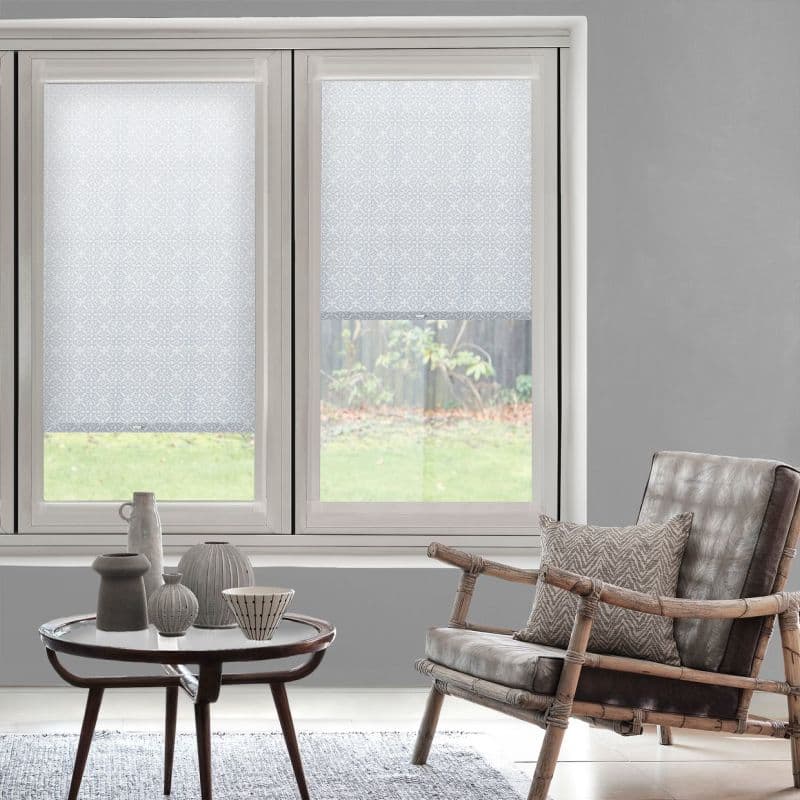 Patterned white roller blinds, covering two adjacent windows (left fully lowered, right partially raised), diffusing daylight into a neutral living room with a rattan-frame leather chair and coffee table.