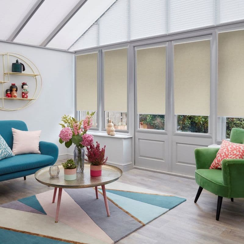 Beige fabric roller blinds covering tall French windows and triangular skylights, lowered to mid-height and filtering daylight; bright conservatory-style living room with colourful chairs, rug, and coffee table.