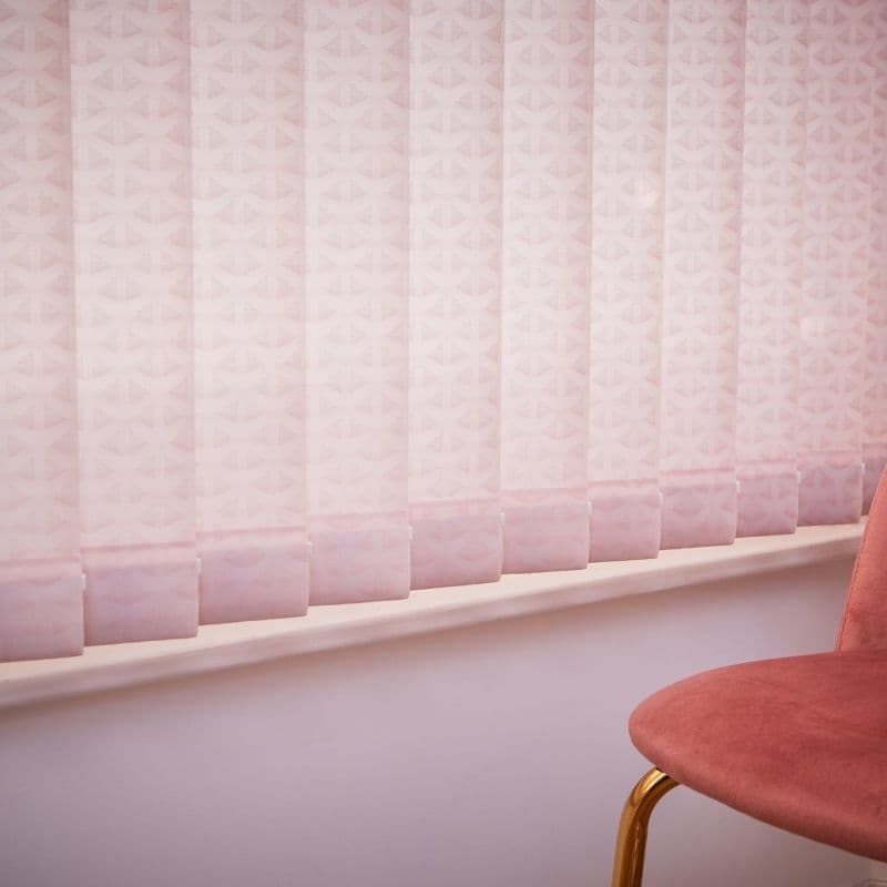 Pink patterned vertical blinds (fabric), fully closed and softly filtering daylight; mounted over a windowsill. Context: pale pink interior wall and a pink velvet chair with gold metal leg visible.