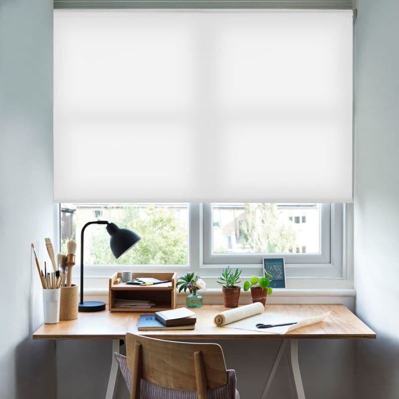 White fabric roller blind, fully lowered across a double window, smooth and semi-opaque, filtering daylight and obscuring most view; above a wooden desk with lamp, brushes and potted plants.