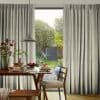 Floor-length pleated beige curtains, ceiling-mounted and drawn apart to reveal a central doorway, filtering soft daylight; dining table with plates, pendant lamp and wooden chairs in a sunny garden-facing room.