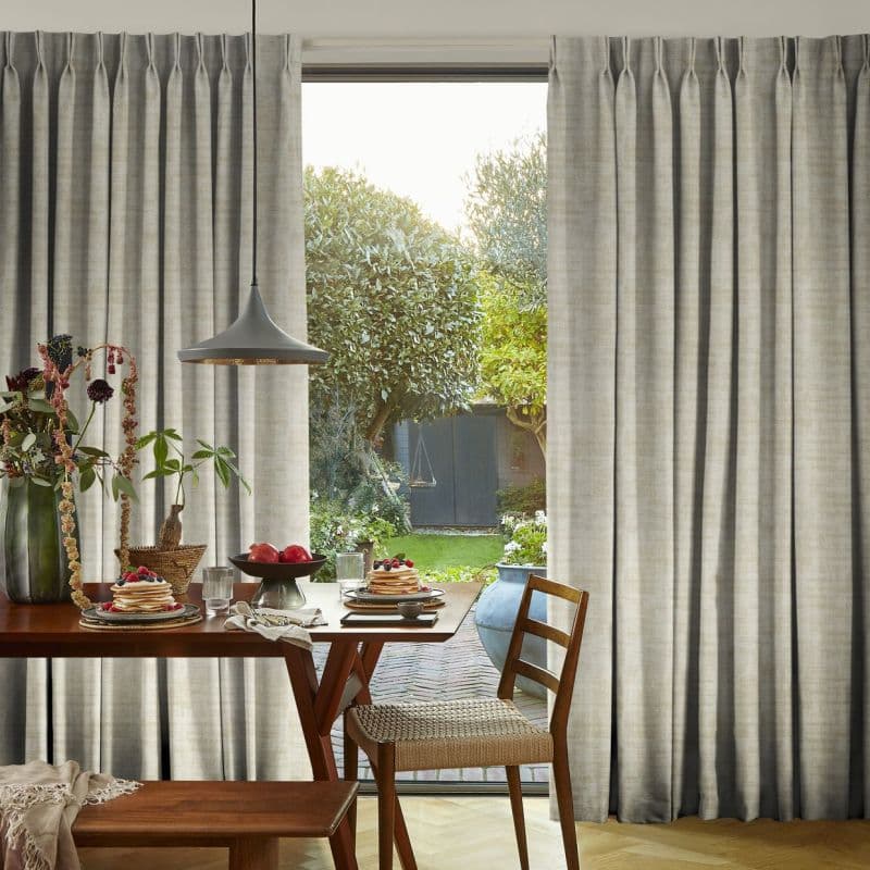 Floor-length pleated beige curtains, ceiling-mounted and drawn apart to reveal a central doorway, filtering soft daylight; dining table with plates, pendant lamp and wooden chairs in a sunny garden-facing room.