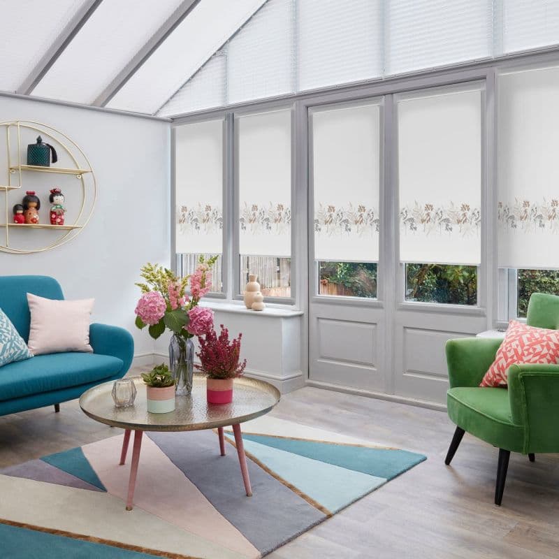 White roller blinds across tall conservatory windows, lowered and closed with a delicate horizontal floral border, filtering bright daylight; cozy sitting room with colorful sofa, green armchair, rug, and flowers.