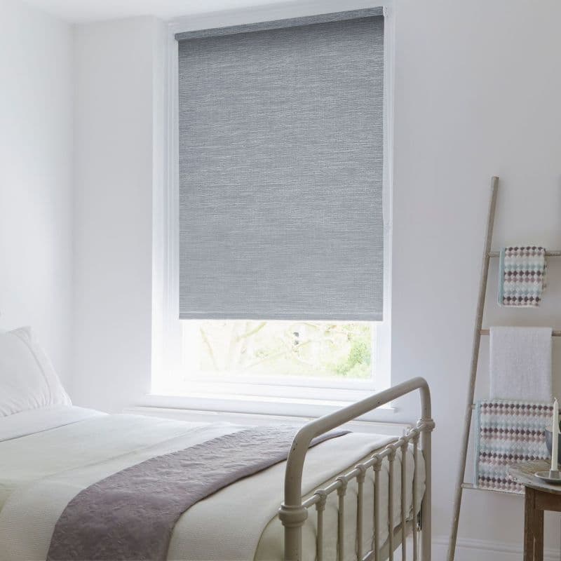 Grey textured roller blind lowered over a single sash window, filtering daylight; in a bright white bedroom with a metal bed frame, soft bedding, and a towel-ladder against the wall.