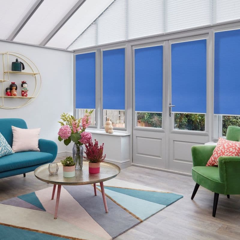 Blue roller blinds covering lower panels of tall French windows and door, lowered; white pleated blinds over upper triangular panes; bright conservatory living room with colorful sofas, rug, coffee table.