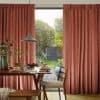 Heavy rust-colored pleated curtains, drawn to frame a sliding door, filtering soft daylight; dining table set for breakfast, wooden chairs in a warm kitchen-dining space overlooking a green garden.