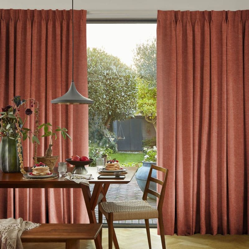Heavy rust-colored pleated curtains, drawn to frame a sliding door, filtering soft daylight; dining table set for breakfast, wooden chairs in a warm kitchen-dining space overlooking a green garden.