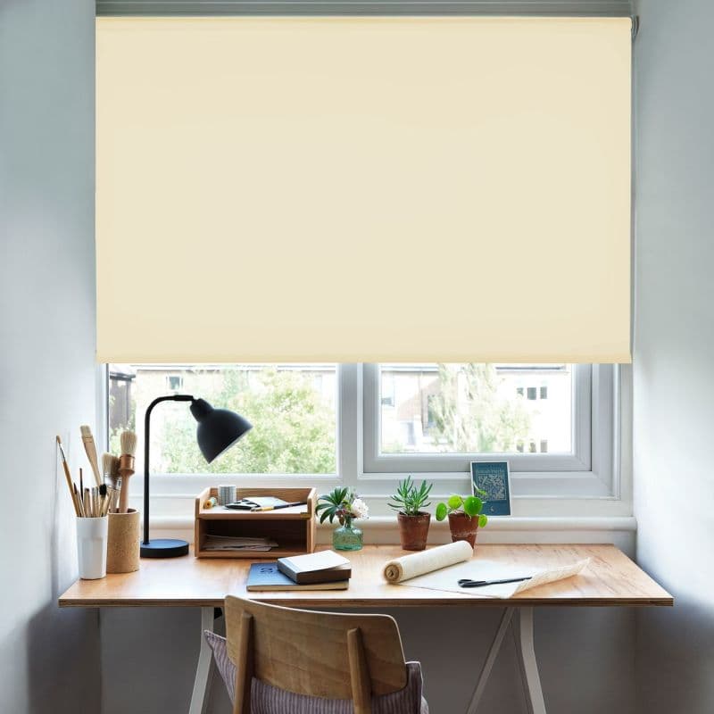 Cream roller blind, lowered almost fully, translucent fabric filtering soft daylight and blocking most of the view; tidy home desk with lamp, brushes, potted succulents, notebooks and wooden chair.