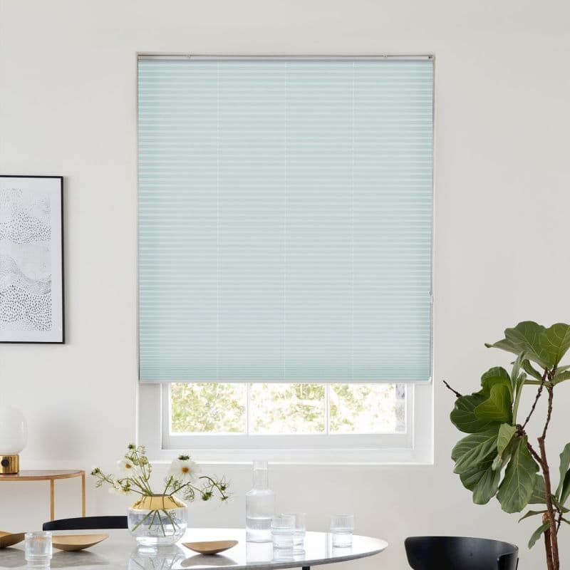 Pale blue pleated blind, fully lowered and filtering light, fitted inside a recessed window; room shows a dining table with glassware and vase, neutral walls, and a fiddle‑leaf fig plant.