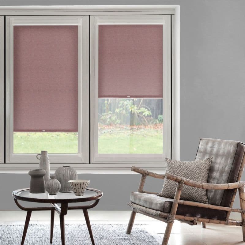 Roller fabric blinds (dusty rose), fitted inside white window frames, partially lowered filtering daylight; living room with gray walls, rattan armchair, patterned cushion and round table with vases, garden outside.