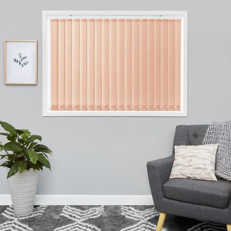 Peach textured vertical blinds in a window frame, closed and diffusing light; in a gray living room with a gray armchair, patterned rug, potted plant, and framed botanical print.