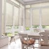 Beige horizontal Venetian blinds covering tall French windows and doors, lowered and filtering soft daylight; bright, sunlit dining nook with white wooden table, chairs, floral cushions, and garden view.
