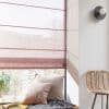 Sheer mauve Roman blind lowered over a window, horizontal folds filtering soft daylight; a window seat with pillows, nearby plant, woven chair and round wall sconce in a bright, cozy corner.