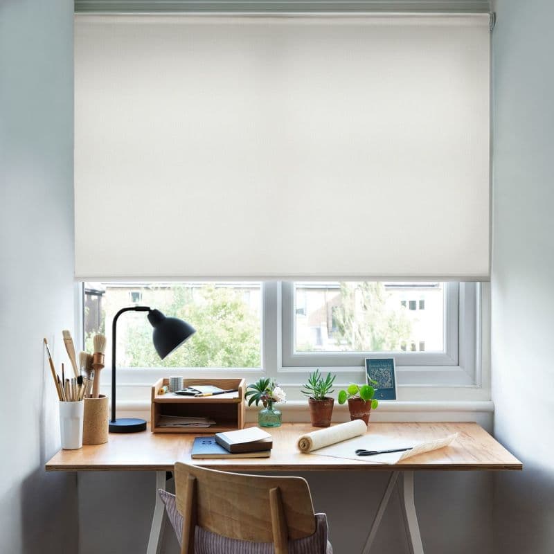 White fabric roller blind, lowered almost fully across a double window with a slim cassette; filtering soft daylight into a small home study with wooden desk, lamp, plants, stationery.