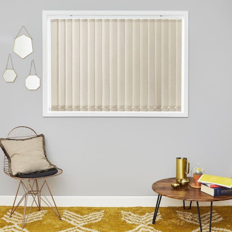 Beige vertical blinds (closed) covering a rectangular window, slats linked by a bead chain at the bottom, filtering light in a living area with chair, wooden table and mustard rug.