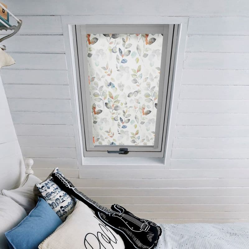Floral roller blind with watercolor leaf pattern, fully lowered, diffusing daylight across a white-painted attic bedroom; slanted shiplap walls, bed with cushions and guitar-shaped pillow below.