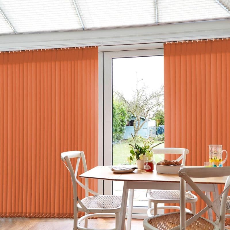 Orange vertical blinds, floor-to-ceiling on a sliding glass door, mostly closed with a central gap, filtering daylight; small bright dining area with wooden table, white chairs, vase and garden view.