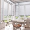 Light-gray horizontal Venetian blinds, fitted inside tall bay windows, partially lowered with closed slats filtering daylight; bright white dining nook with wooden table and chairs, garden visible beyond.